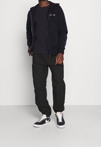 Sudadera con cremallera color navy con franjas laterales rojas, camiseta negra, pantalones cargo negros y zapatillas negras con detalles blancos. Texturas de tela suaves.