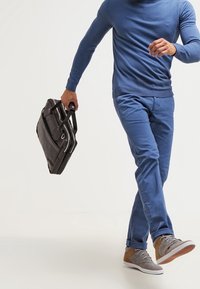 Schwarze Lederaktentasche, hellblauses langärmliges Hemd und hellblaue Chinos mit grauen und beigen Sneakers. Das Outfit zeichnet sich durch ein lässiges, modernes Design aus.