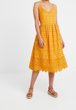 Żółta sukienka bez rękawów z dekoltem w serek i haftowanym ażurowym wzorem, z rozkloszowanym dołem i falowanym brzegiem. Połączona z brązowymi espadrylami.