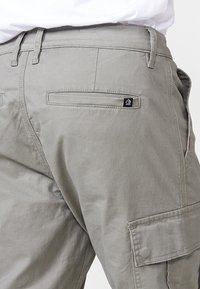 Pantalones cargo grises con bolsillos laterales, bolsillo trasero con logo, trabillas para cinturón y una textura suave. El corte incluye un ligero estrechamiento hacia las piernas.