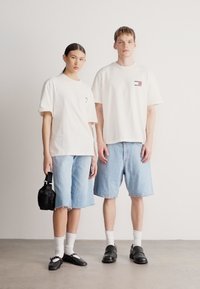 T-shirts brancas de algodão, calções de ganga azul claro, mala preta, sapatos pretos, meias brancas, em pé num cenário minimalista.