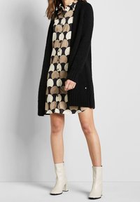 Czarny sweter na drutach narzucony na sukienkę w wzór z beżowymi i brązowymi okręgami. Dopełnieniem stroju są białe botki na słupku.