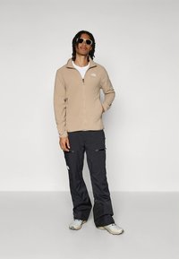 Beige Fleecejacke mit Reißverschluss über einem weißen T-Shirt, kombiniert mit schwarzen Hosen mit Reißverschlusstaschen und grauen Wanderschuhen. Sonnenbrille getragen.