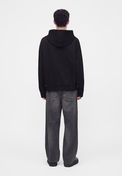 Homme vu de dos portant un sweat à capuche noir, un jean gris ample et des chaussures noires sur un fond blanc uni.