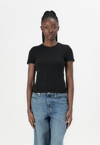 Black short-sleeve top featuring a textured pattern of small silver dots. Paired with loose blue jeans. Neat, fitted silhouette.