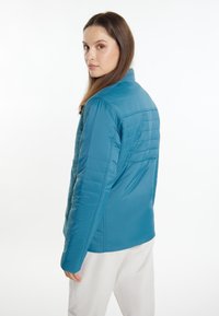 Giacca leggera in blu, tipo puffer, con colletto strutturato, dettagli di cuciture orizzontali e tasche laterali con zip; texture liscia e vestibilità affusolata.