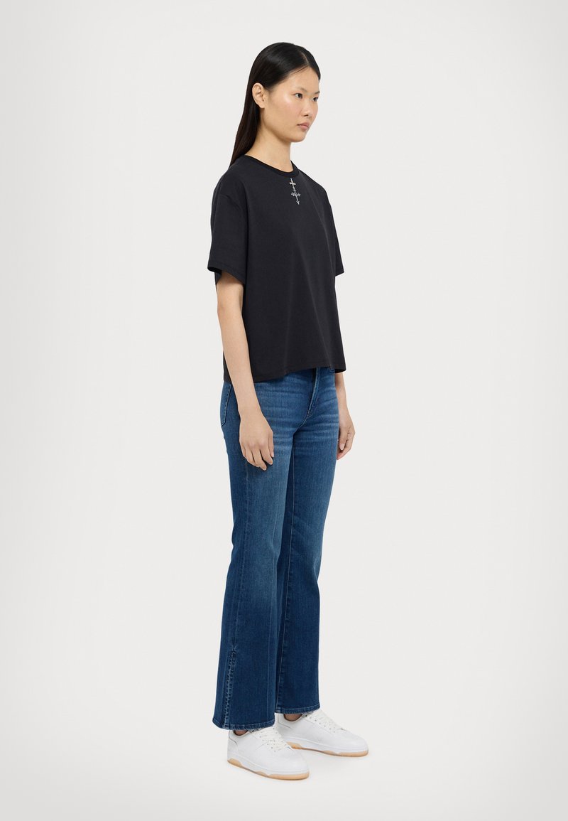 Sorte cropped t-shirt med rund hals og sølv korsdetaljer, parret med flagrende blå denimjeans og hvide sneakers.