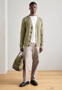 Zelený rebrovaný cardigan s čiernymi gombíkmi, nosený cez biele tričko. Béžové nohavice a čierne čižmy. Drží zelenú tašku s hnedými akcentmi.