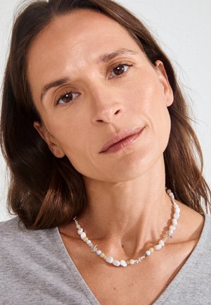 Vrouw met bruin haar die een grijze top en een witte gekralen ketting met kleine bloemvormige accenten draagt, neutrale gezichtsuitdrukking, lichte achtergrond.