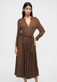 Καφέ πουά φόρεμα με μακριά μανίκια, V-λαιμόκοψη και ζώνη στη μέση. Διαθέτει αέρινη φούστα και απαλή υφή υφάσματος.