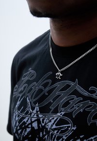 Colier din argint cu un accent negru în formă de inimă, purtat de o persoană îmbrăcată cu un tricou grafic negru, decorat cu litere și un design gri.