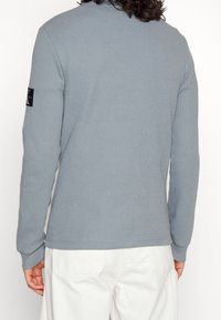 Langärmliges, hellgraues Waffelstrickshirt mit gerippten Bündchen und einem schwarzen Logopatch am oberen Arm, strukturiertem Stoff und klaren Linien.