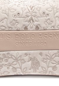 Beige tygpås med intrikata blommönster och ett texturerat beige band med "U.S. POLO ASSN. SINCE 1890" präglat.