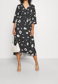 Svart omlottklänning med vitt blommönster, trekvartsärmar, knytning i midjan och en svävande nederdel. Bärs med sandaletter med remmar och en lätt handväska.