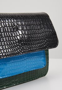 Kolme pinottua suorakaiteenmuotoista käsilaukkua, joissa on kiiltävä krokotiilinkuosi mustana, kirkkaan sinisenä ja tummanvihreänä valkoisella pinnalla.