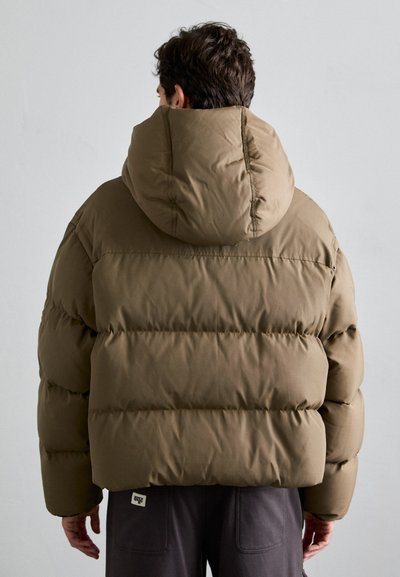 Olivgrüne Pufferjacke mit Kapuze, die ein kastiges, gestepptes Design und eine weiche Textur aufweist. Auffällige sichtbare Nähte und ein Logo-Etikett.