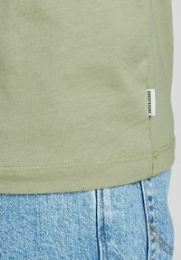 Oljenvihreä puuvillapaita suoran leikkauksen kanssa, jossa on sivutägi. Yhdistetty vaaleansiinisiin farkkushortseihin, joissa on näkyvät ompelusaumat.