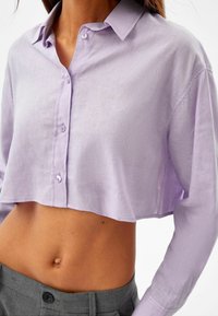 Cropped button-up shirt in light lavender fabric, featuring long sleeves, a collar, and a slightly textured finish. Buttons are matching color.