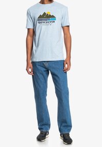 Hombre con camiseta Quiksilver de color azul claro con diseño de montaña y ola, jeans azules y zapatillas negras, de pie contra un fondo blanco.