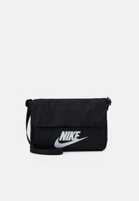Melnas auduma sūtījumu soma ar atloku, kurai ir izteikta sudraba Nike zīme priekšpusē un regulējama siksna.