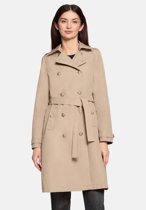 Frau trägt einen beigen, doppelt geknöpften Trenchcoat mit Taillengürtel und dekorativen Knöpfen, steht vor einem schlichten hellen Hintergrund.