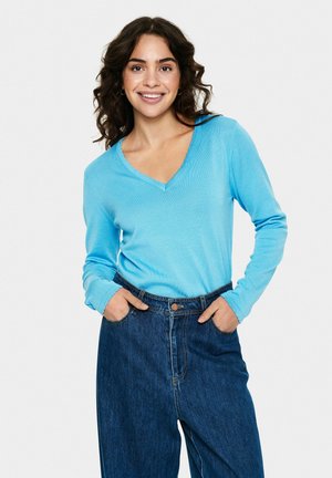 Ljusblå långärmad V-ringad topp i mjukt tyg, kombinerad med högmidjade mörka denimjeans med fickor och knappstängning.