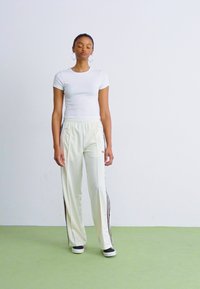 Weißes, eng anliegendes T-Shirt, cremefarbene Jogginghose mit schwarzen Seitenstreifen und schwarze Turnschuhe. Einheitliche Farben, lockere Passform, minimalistisches Design.