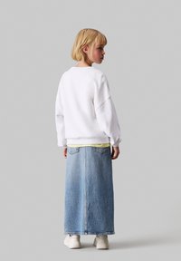 Biele oversized mikina, žltá vrstva pod ňou, skombinovaná s dlhou modrou džínovou sukňou. Biele tenisky dopĺňajú outfit.