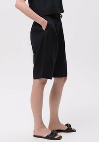 Zwarte shorts van zacht materiaal, met zijzakken, een ontspannen pasvorm en een subtiele glans. Gekoeld met zwarte geknoopte slidesandalen.