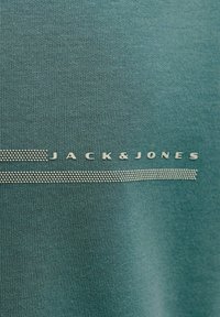 Chemise turquoise à manches courtes en tissu texturé, ornée d'un logo argenté "JACK&JONES" et d'un accent horizontal en points.