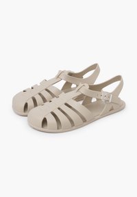Sandalias de plástico beige con diseño de punta abierta, cortes verticales, correa ajustable en el tobillo con hebilla y suela plana para mayor comodidad.