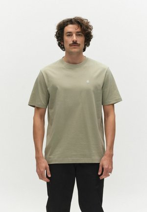 Mann mit lockigem Haar und Schnurrbart, trägt ein hellolivgrünes Kurzarm-T-Shirt und schwarze Hose, steht vor einem schlichten weißen Hintergrund.