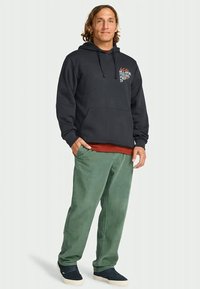 Man in een zwarte hoodie met een voorzak en logo, groene corduroy broek en zwarte sneakers, staand met één hand in de zak.