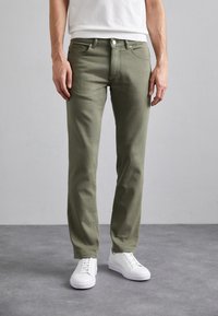 Oliwkowe spodnie z bawełny w kroju slim fit, z pięcioma kieszeniami oraz z przodem na guziki, w zestawieniu z białymi trampkami.