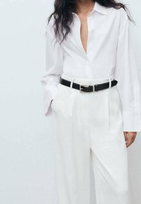 Chemise blanche à manches longues avec un col en V profond, associée à un pantalon blanc taille haute et une ceinture en cuir noire avec une boucle en argent.