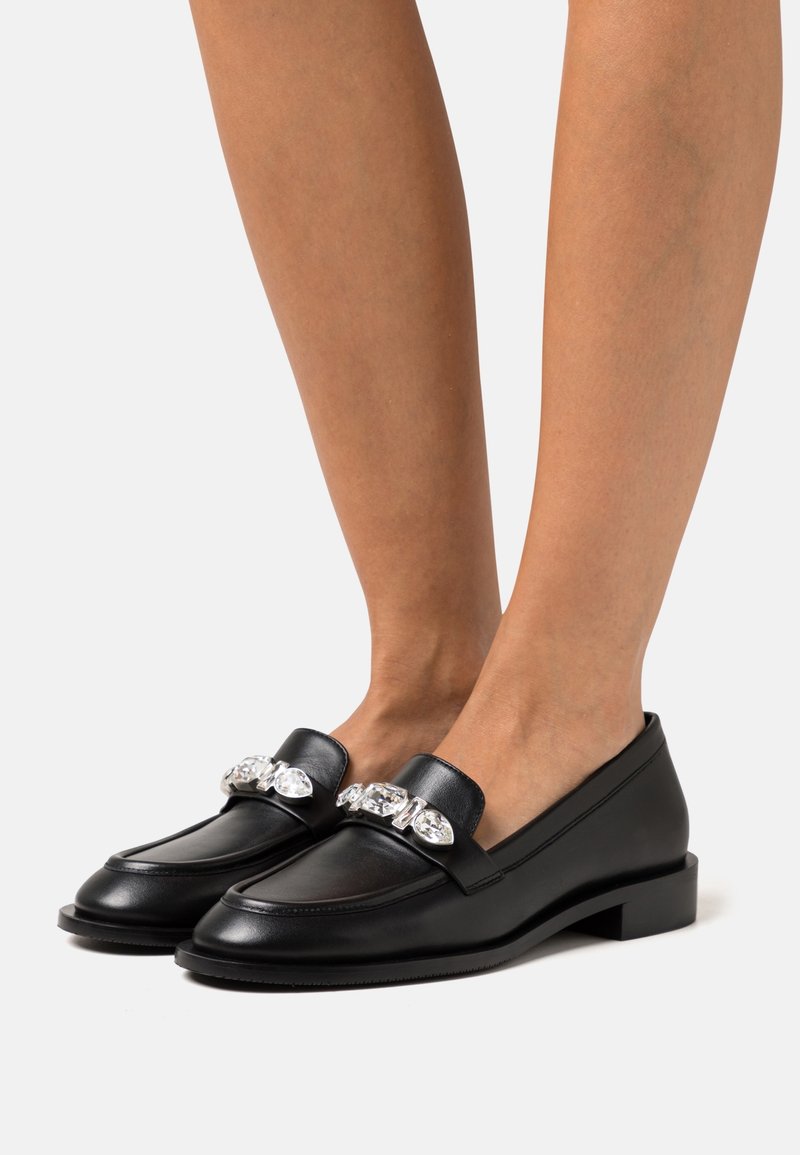 Mocassins en cuir noir avec de grandes garnitures en pierres précieuses transparentes portés pieds nus sur un fond clair uni.