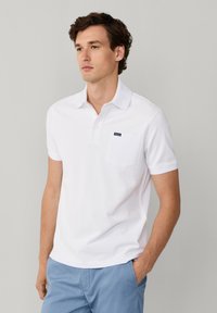 Jeune homme portant un polo blanc à manches courtes avec une poche poitrine et un pantalon bleu clair, debout devant un fond uni gris clair.