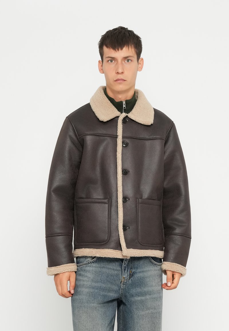 Donkerbruine leren jas met een beige schapenvacht kraag en afwerking, voorzien van twee voorzakken en knoopsluiting. Klassiek boxy ontwerp.
