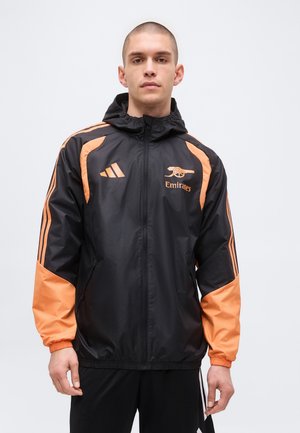Giovane uomo che indossa una giacca con cappuccio nera e arancione con cerniera e con i loghi del club di calcio Arsenal e Adidas sul davanti.