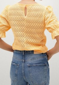Blusa amarilla de ojo de perdiz con mangas abullonadas y detalle de ojal en la parte posterior, combinada con jeans de mezclilla azul de tiro alto que tienen bolsillos traseros visibles.
