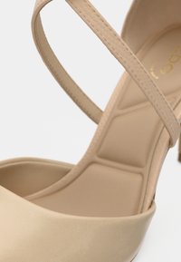 Gros plan sur une chaussure à talon beige avec des lanières fines et une semelle intérieure rembourrée, montrant les coutures détaillées et la texture.