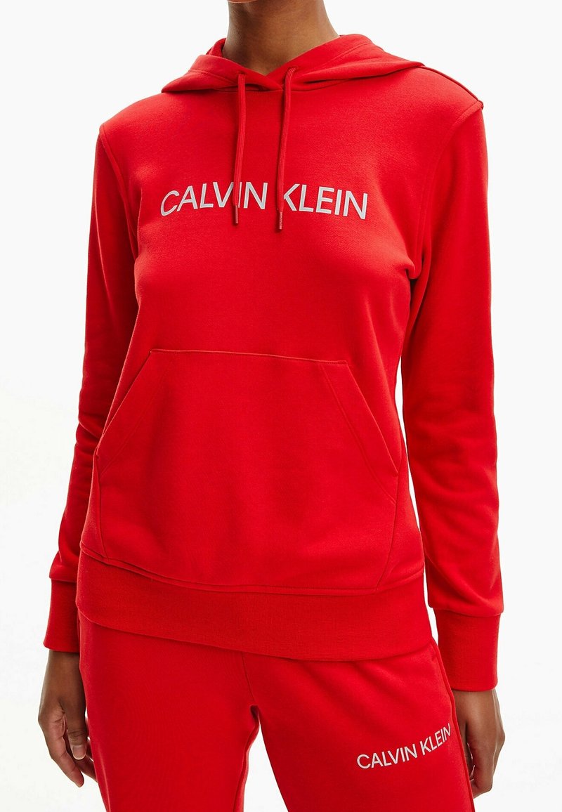 Hoodie rouge avec une poche avant, capuche à cordon de serrage, et logo "CALVIN KLEIN" blanc. Fabriqué en tissu doux et texturé, avec des poignets et un ourlet côtelés.