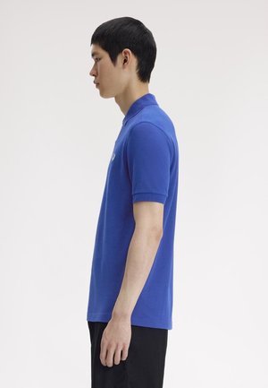 Jonge man staand in profiel, gekleed in een blauw poloshirt met korte mouwen en zwarte broek, tegen een effen achtergrond.