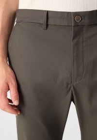 Donkergrijze pantalon met een soepele textuur, voorzien van een knoopsluiting en subtiele stiksels, getoond met een hand nabij de tailleband.