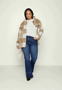 Casaco quadrados em bege e branco, com grandes botões, combinado com uma blusa branca e jeans azuis flare. Tecido texturizado e bolsa acolchoada.