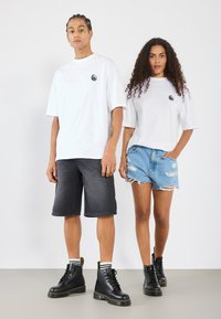 Hvide oversized t-shirts med sort logo, parret med afslappede denimshorts og sorte snørebåndsstøvler med gule accenter. Glatte, afslappede materialer.