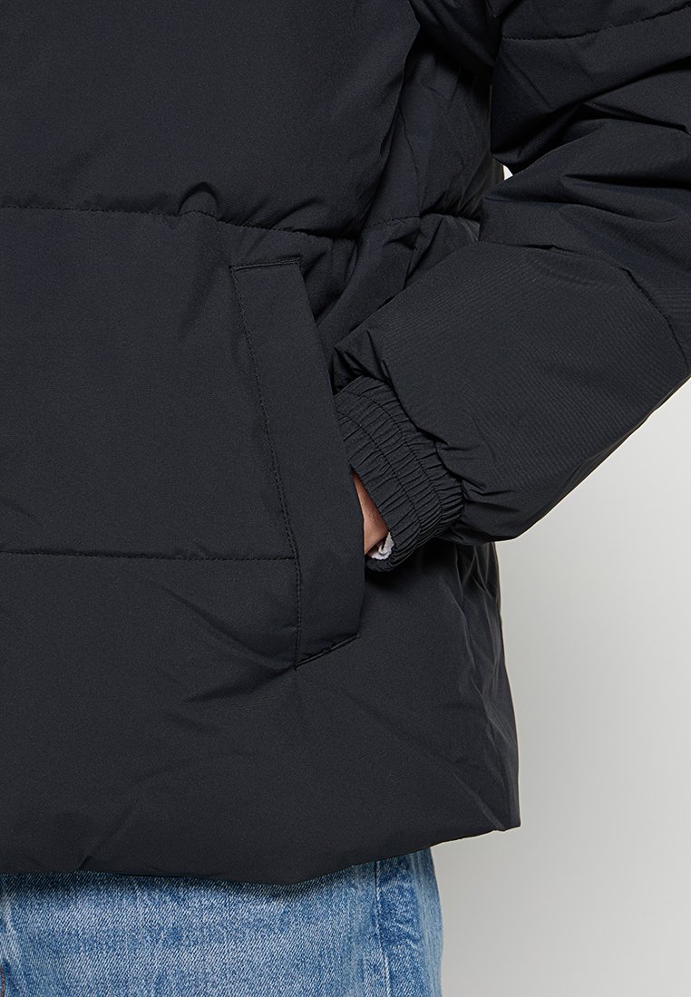 Chaqueta acolchada negra con puños elásticos, un bolsillo frontal y una textura suave, combinada con jeans de mezclilla azules.