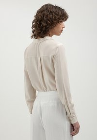 Donna con capelli ricci che indossa una blusa lunga a maniche, di color crema, infilata in pantaloni bianchi a vita alta, rivolta di lato contro uno sfondo semplice.