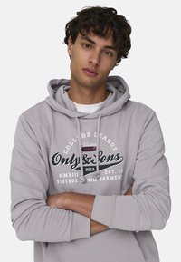 Sudadera gris con un gráfico en la parte frontal que incluye texto, cordones y un bolsillo tipo canguro. Tejido suave con un corte holgado y puños acanalados.