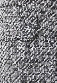 Tissu en tweed noir et blanc avec des éclats d'or, présentant un motif texturé. Un bouton circulaire en argent est ajouté, apportant une touche de détail.